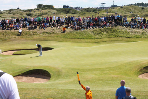 Golfer on a green golf course with a crowd watching, including a person in an orange jacket waving.